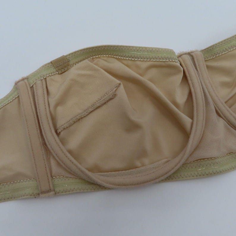 May include: A beige strapless bra with a smooth, seamless design. The bra features a structured underwire and a soft, neutral fabric. The interior lining is a matching beige color, and the edges are trimmed with a delicate, light green stitching.