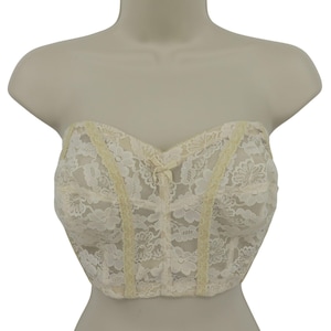 Pode incluir: Bustier sem alças branco sujo com sobreposição de renda floral. O bustier apresenta um decote em coração, um pequeno laço no centro e acabamento vertical amarelo. A peça é exibida em um manequim.