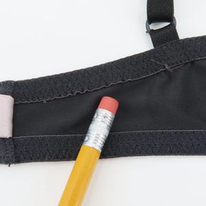 Op de afbeelding: Close-up van een zwarte bh met verstelbare bandjes en een haaksluiting. De bh heeft een gladde, matte stof met een getextureerde rand. Een gele potlood met een roze gum ligt op de bh.