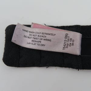 Op de afbeelding: Een close-up van een zwart kledingstuk met een roze en wit label. Het label bevat wasvoorschriften: "HANDWAS KOUD APART, NIET BLEKEN, NIET WRINGEN, HERVORMEN, PLAT LIGGEN OM TE DROGEN." Het label bevat ook de nummers "154212-093, RN# 70817, ID# 106, 08 03."