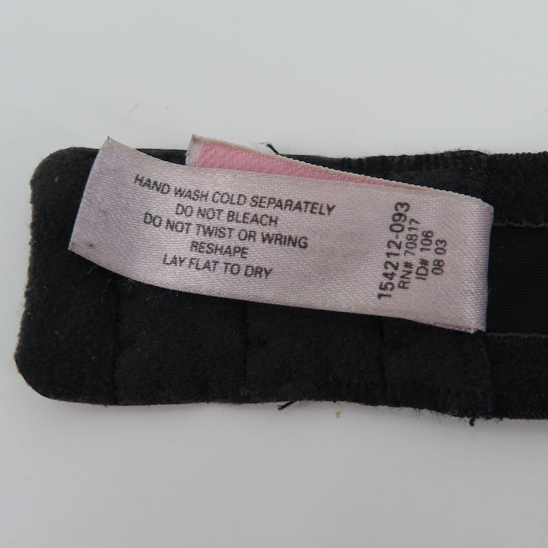 Op de afbeelding: Een close-up van een zwart kledingstuk met een roze en wit label. Het label bevat wasvoorschriften: "HANDWAS KOUD APART, NIET BLEKEN, NIET WRINGEN, HERVORMEN, PLAT LIGGEN OM TE DROGEN." Het label bevat ook de nummers "154212-093, RN# 70817, ID# 106, 08 03."