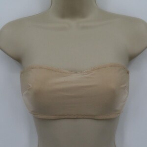 May include: A beige strapless bra displayed on a mannequin. The bra has a smooth, seamless design with a curved neckline and a stitched hem. The neutral color makes it versatile for various outfits.
