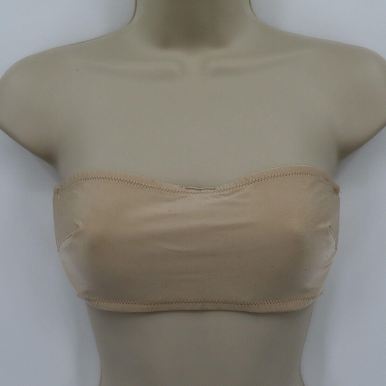 May include: A beige strapless bra displayed on a mannequin. The bra has a smooth, seamless design with a curved neckline and a stitched hem. The neutral color makes it versatile for various outfits.