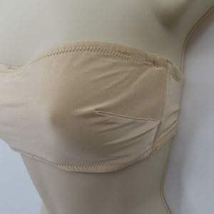 May include: A beige strapless bra with a smooth, seamless design. The bra features a subtle, neutral color and is constructed with a soft fabric. The edges are finished with a delicate, stitched trim, and the cups are lightly padded.