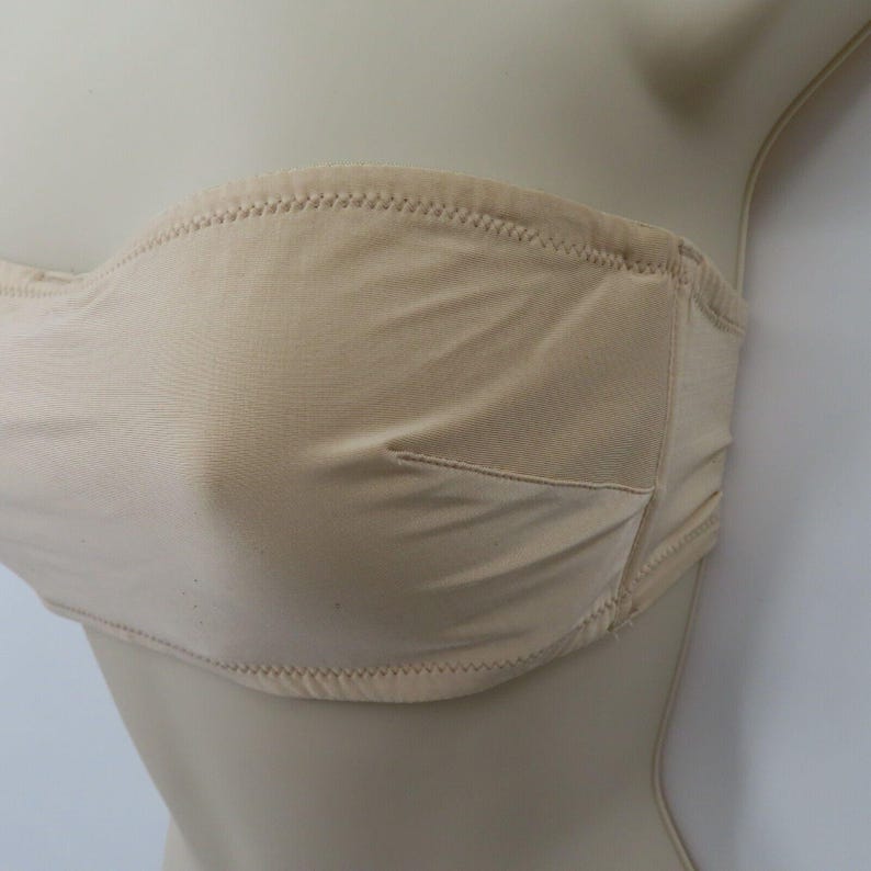 May include: A beige strapless bra with a smooth, seamless design. The bra features a subtle, neutral color and is constructed with a soft fabric. The edges are finished with a delicate, stitched trim, and the cups are lightly padded.