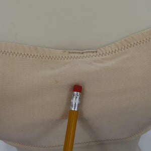 May include: Close-up of a beige undergarment with a zigzag stitched edge. A yellow pencil with a red eraser rests on the fabric, providing a size reference. The undergarment appears to be made of a soft, smooth material.