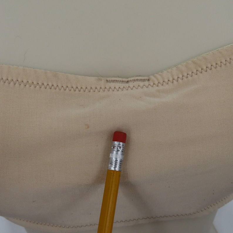 May include: Close-up of a beige undergarment with a zigzag stitched edge. A yellow pencil with a red eraser rests on the fabric, providing a size reference. The undergarment appears to be made of a soft, smooth material.