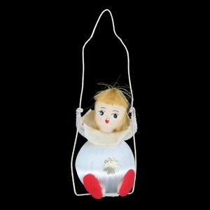 Peut inclure: Ornement de Noël vintage représentant une poupée dans une balançoire. La poupée a un visage peint avec des yeux bleus et des lèvres rouges, des cheveux blonds et des pieds rouges. La poupée porte une robe blanche avec un col crème et est suspendue par une balançoire en métal blanc.