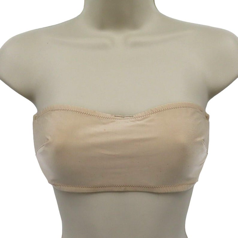 May include: A beige strapless bra displayed on a mannequin. The bra is made of a smooth fabric and features a scalloped top edge and a stitched hemline. The bra is designed to provide support without straps.