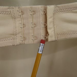 May include: Close-up of a beige bra clasp with three rows of hooks and eyes. The fabric is a light beige color, and the stitching is visible. A yellow pencil with a red eraser points to the clasp.