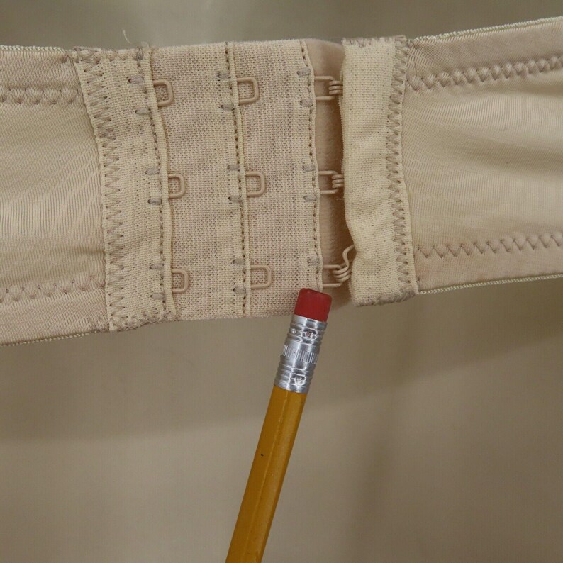 May include: Close-up of a beige bra clasp with three rows of hooks and eyes. The fabric is a light beige color, and the stitching is visible. A yellow pencil with a red eraser points to the clasp.