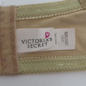 May include: Close-up of a beige bra with a tag that reads "VICTORIA'S SECRET" and a heart logo. The tag also indicates a size of 34C. The bra has a light green trim and a beige lining.