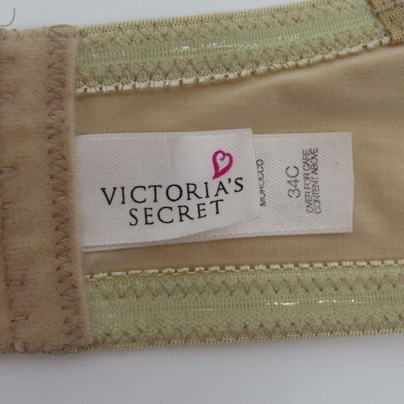 May include: Close-up of a beige bra with a tag that reads "VICTORIA'S SECRET" and a heart logo. The tag also indicates a size of 34C. The bra has a light green trim and a beige lining.