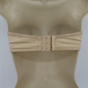 May include: A beige strapless bra with a hook-and-eye closure. The bra is made of a smooth fabric and features a bandeau style. The closure has three sets of hooks for adjustable sizing. The bra is displayed on a mannequin.