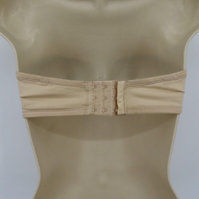 May include: A beige strapless bra with a hook-and-eye closure. The bra is made of a smooth fabric and features a bandeau style. The closure has three sets of hooks for adjustable sizing. The bra is displayed on a mannequin.