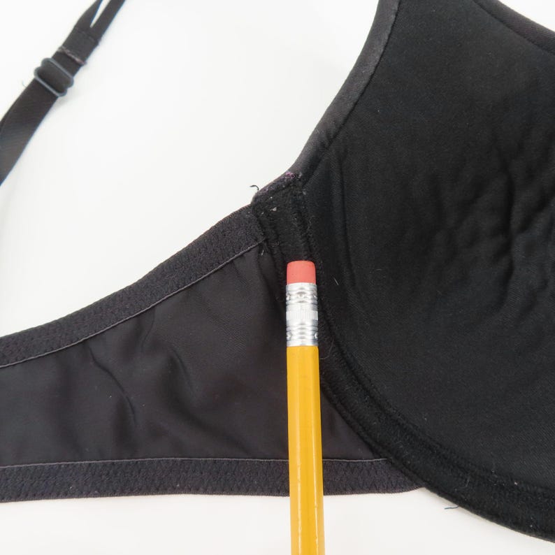 Op de afbeelding: Close-up van een zwarte bh met verstelbare bandjes. De bh is gemaakt van een glad, effen zwart materiaal. Een gele potlood met een roze gum ligt naast de bh.
