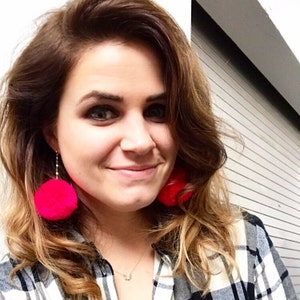 May include: A woman with long brown hair wearing a black and white plaid shirt and large pink pom-pom earrings.