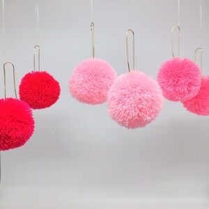 May include: Five fluffy pom-pom ornaments hanging from gold chains. Three are pink and two are red.