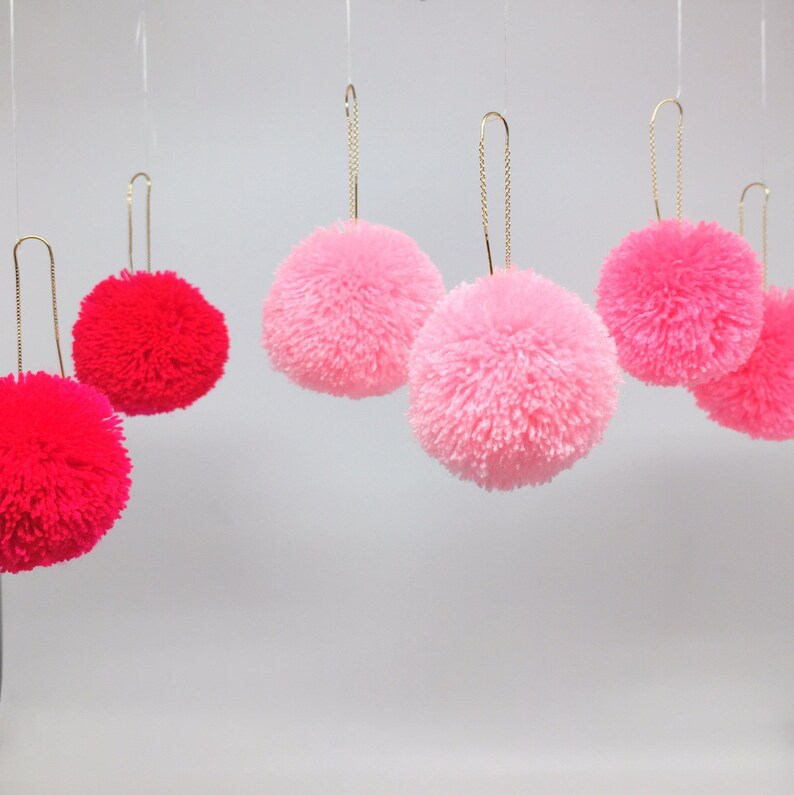 May include: Five fluffy pom-pom ornaments hanging from gold chains. Three are pink and two are red.