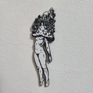 May include: A black and white enamel pin featuring a stylized nude figure with a mushroom cap headpiece. The pin has a detailed design with a glossy finish, perfect for jackets, bags, or hats. The pin is a unique accessory.