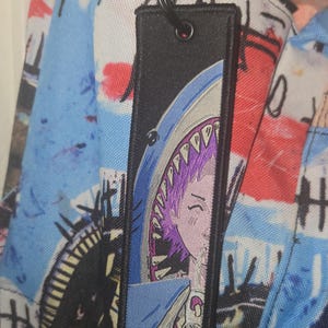 May include: Black rectangular tag with a shark-themed anime design. The tag features a shark's open mouth with a person's face inside. The tag is attached to a backpack with a colorful graffiti-style pattern.