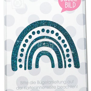 Peut inclure: Un autocollant thermocollant bleu turquoise représentant un arc-en-ciel avec des paillettes et des pois blancs. L'autocollant est emballé sur une carte blanche avec le texte "BÜGEL-BILD" et "MIMMIS TRAKTOR®".