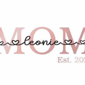 Könnte beinhalten: Rosa-weißes Grafik mit dem Wort "MOM" in großen Buchstaben. Das Wort "Leonie" ist in Kursivschrift mit kleinen Herzen auf beiden Seiten geschrieben. Der Text "Est. 2022" befindet sich unter der Grafik.