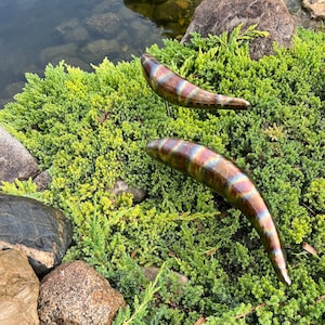 May include: Two metal fish sculptures with iridescent blue, green, and brown colors are placed in a bed of green foliage. The fish are positioned with their tails pointing up.