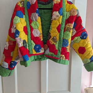 May include: A quilted jacket constructed from multicolored hexagonal fabric patches in shades of red, yellow, blue, and green. The garment features a wide shawl collar, long sleeves with green cuffs, and a single large brown button closure.