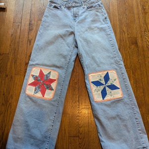 May include: Light blue denim jeans featuring wide legs and a high waist. Each knee area is decorated with a square fabric patch displaying a multi-pointed star pattern. The left patch uses red and floral fabric, while the right uses blue and floral fabric.