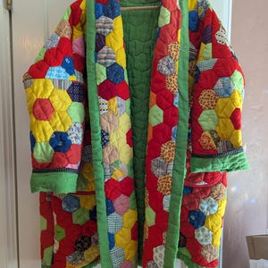 May include: A long-sleeved quilted jacket constructed from a patchwork of multicolored hexagonal fabric pieces. The garment features a vibrant array of red, yellow, blue, and patterned textiles, finished with solid green trim along the front opening and cuffs.