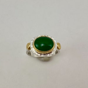 Può includere: Un anello d'argento con una pietra preziosa verde incastonata in una ghiera d'oro. L'anello ha una fascia testurizzata e dettagli dorati.