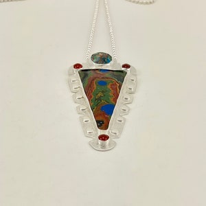 May include: A silver necklace with a triangular pendant featuring a colourful, abstract design. The pendant is surrounded by a silver frame with small, decorative details. The necklace is adorned with small, silver beads.
