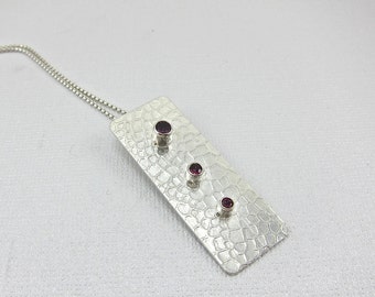 May include: A silver pendant necklace with a textured rectangular charm featuring three small purple gemstones.