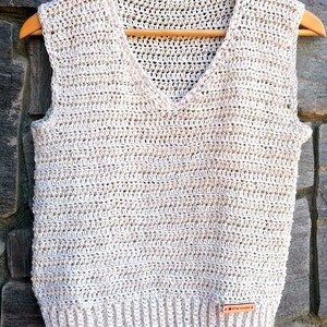Puede incluir: Un chaleco sin mangas de ganchillo en color crema con cuello en V. El chaleco tiene una pequeña etiqueta de cuero marrón con el texto "DIY Crochet" en ella.