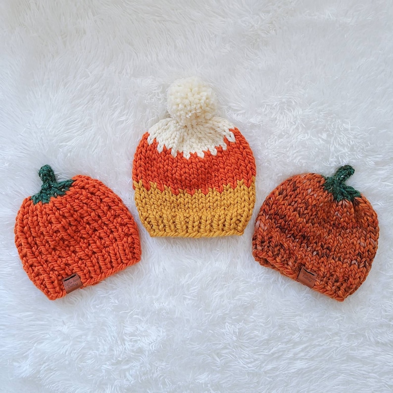 May include: Three knitted hats in the shape of pumpkins and candy corn. The hats are orange, yellow and white.