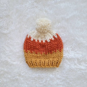 May include: A knitted hat with a white pom pom on top. The hat is striped with orange, white, and yellow.
