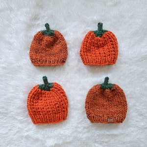 May include: Four knitted pumpkin hats in orange and green. The hats are arranged in a square pattern on a white fluffy surface.