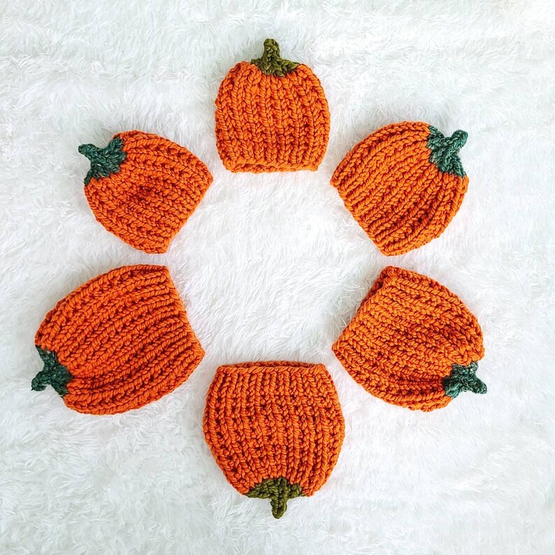 May include: Six orange knitted pumpkin hats with green stems. The hats are arranged in a circle on a white fluffy surface.