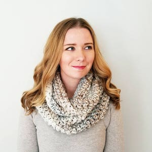 May include: A woman wearing a gray sweater and a crocheted infinity scarf with a gradient of beige, white, and gray.