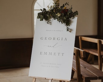 May include: A white sign with the text "Welcome to the wedding of GEORGIA and EMMETT 17 SEPTEMBER 2022 Immerse in the Terra Valley" on an easel with greenery on top.