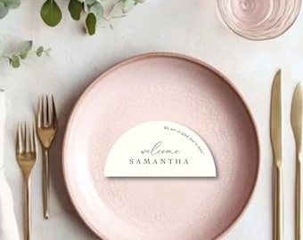 May include: A table setting with a pink plate, gold utensils, and a glass of water. A folded linen napkin and sprigs of greenery are also present. A welcome card with the name "Samantha" is on the plate.