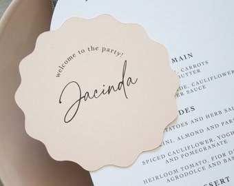 Könnte beinhalten: Eine blassrosa, wellenförmige, kreisförmige Platzkarte mit dem schwarz gedruckten Text "welcome to the party!" und dem Namen "Jacinda" in Schreibschrift, die auf einer weißen Menükarte mit schwarzem Text und einem hellrosa Teller liegt.