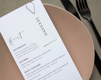 May include: A white menu card with the word "Feast" in elegant script, listing courses like Antipasto and Entrée. A tag with "Luciano" is attached with a silver paper clip. The menu rests on a pink plate, with a black fork and knife.