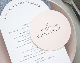 May include: A pink round welcome sign with the text "welcome Christina" in black script font.
