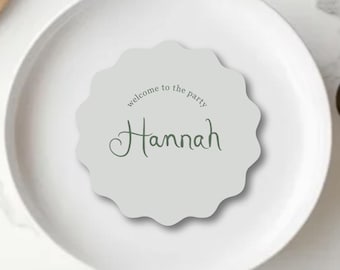 Könnte beinhalten: Eine hellgraue Tischkarte mit gewelltem Rand, auf der der Name "Hannah" in Schreibschrift und der Text "welcome to the party" steht. Die Karte liegt auf einem weißen Teller, daneben liegen ein Löffel und ein Messer.