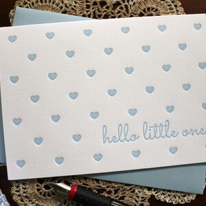 May include: A white card with a blue background and a blue envelope. The card has a pattern of blue hearts and the text "hello little one".