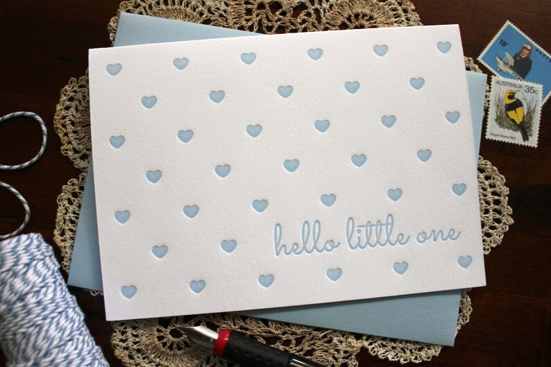 May include: A white card with a blue background and a blue envelope. The card has a pattern of blue hearts and the text "hello little one".