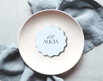 Könnte beinhalten: Eine hellblaue Platzkarte mit gewelltem Rand und dem Text "hello ALICIA" in schwarzer Schrift liegt auf einem gesprenkelten weißen Teller. Eine blaue Leinen-Serviette ist um den Teller drapiert. Das Bild hat eine weiche, neutrale Ästhetik.