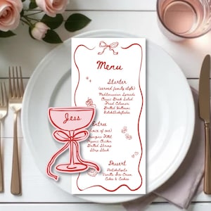 May include: A white menu card with a red border and menu items, including starters, entrees, and desserts. A pink cocktail glass with the name "Jess" is on the left. The menu is on a white plate with gold cutlery and a pink napkin.